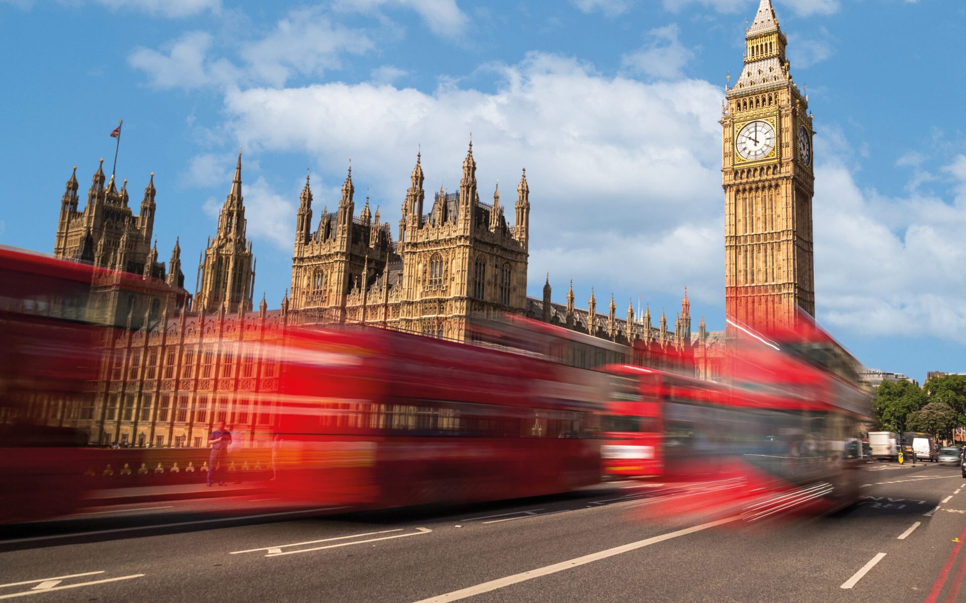 Blick auf den Elizabeth Tower und das Parlamentsgebäude in London unter blauem Himmel; im Vordergrund fahren rote Doppeldeckerbusse mit Bewegungsunschärfe über die Straße.