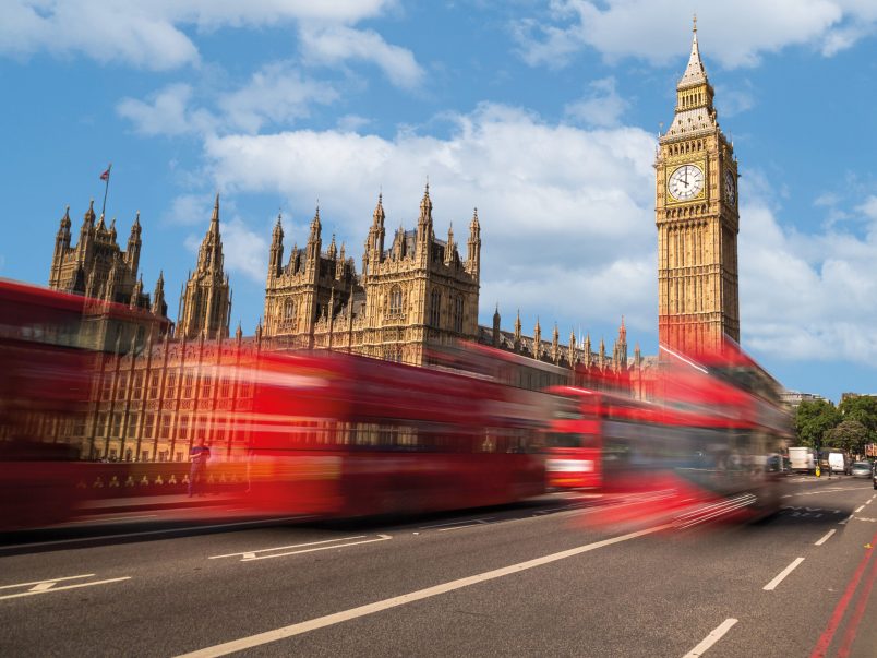 Blick auf den Elizabeth Tower und das Parlamentsgebäude in London unter blauem Himmel; im Vordergrund fahren rote Doppeldeckerbusse mit Bewegungsunschärfe über die Straße.