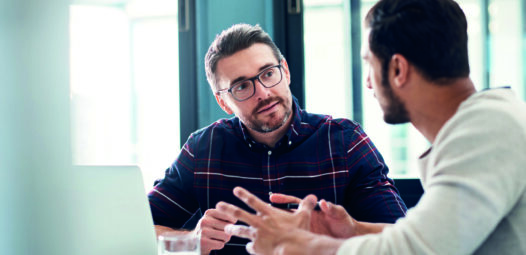 Zwei Männer sitzen an einem Tisch mit Laptop und unterhalten sich in einem hellen Büro; einer erklärt etwas mit Gesten, während der andere aufmerksam zuhört.