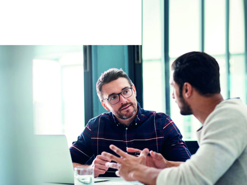 Zwei Männer sitzen an einem Tisch mit Laptop und unterhalten sich in einem hellen Büro; einer erklärt etwas mit Gesten, während der andere aufmerksam zuhört.