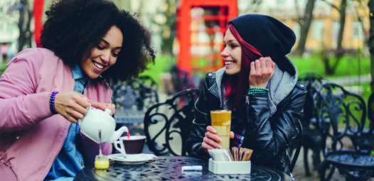 Zwei junge Frauen sitzen an einem Tisch in einem Straßencafé; eine gießt Tee ein, die andere hält ein Latte-Glas. Im Hintergrund steht eine rote Telefonzelle.