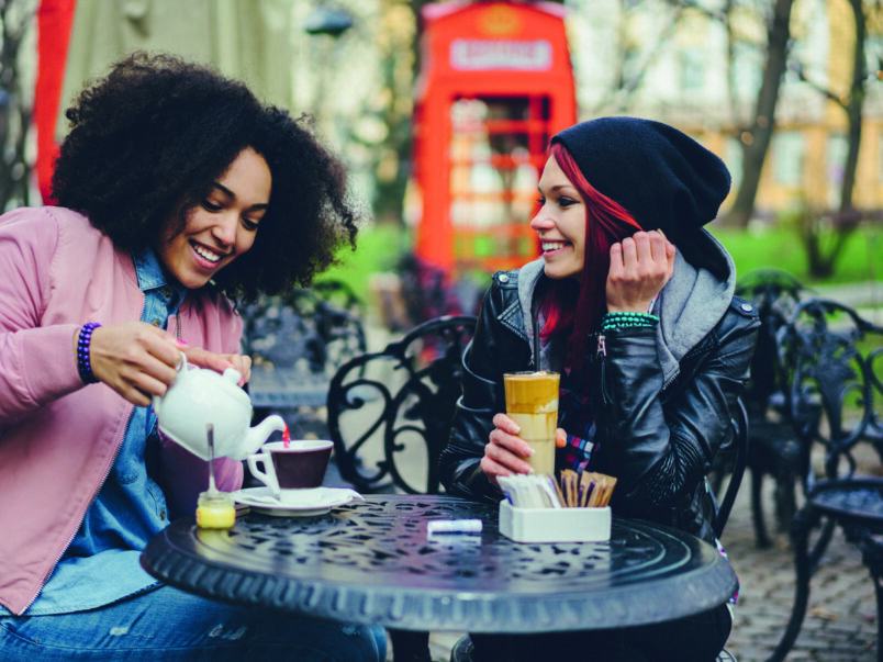 Zwei junge Frauen sitzen an einem Tisch in einem Straßencafé; eine gießt Tee ein, die andere hält ein Latte-Glas. Im Hintergrund steht eine rote Telefonzelle.