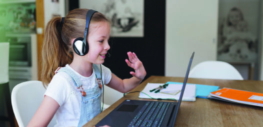 Mädchen mit Headset sitzt an einem Tisch vor einem Laptop, benutzt eine Maus und winkt in die Kamera; neben ihr liegen Hefte und Stifte. Oben im Bild ist viel freie Fläche.