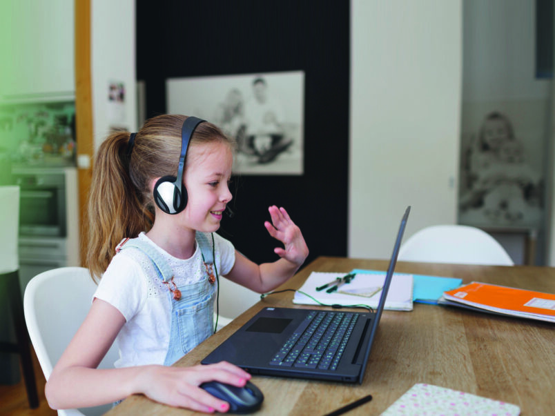 Mädchen mit Headset sitzt an einem Tisch vor einem Laptop, benutzt eine Maus und winkt in die Kamera; neben ihr liegen Hefte und Stifte. Oben im Bild ist viel freie Fläche.