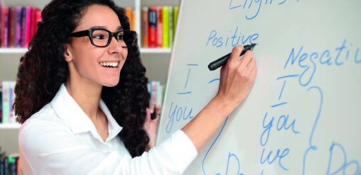 Lächelnde Lehrerin mit Brille schreibt an ein Whiteboard mit englischen Grammatikbeispielen; im Hintergrund stehen Bücherregale.