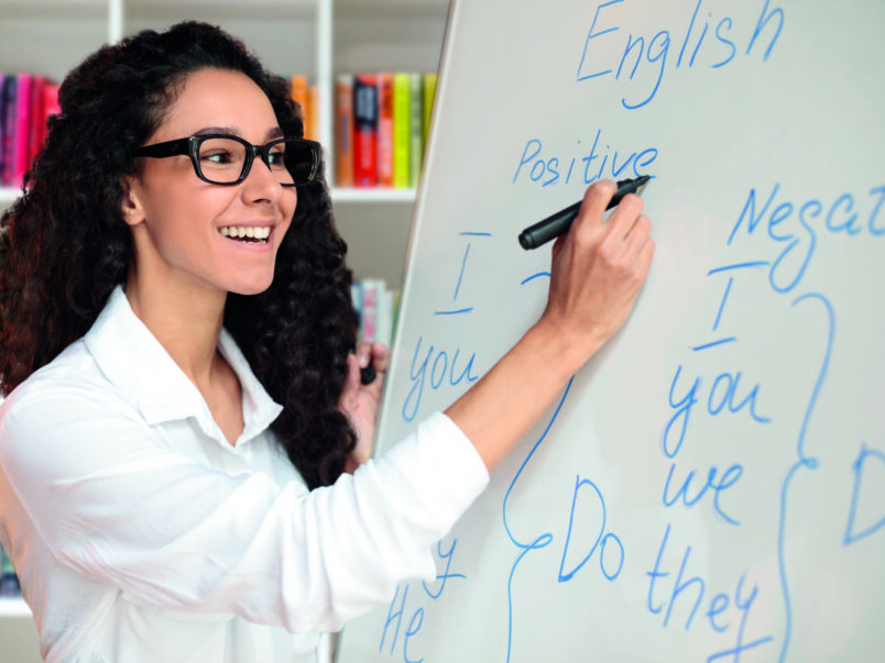 Lächelnde Lehrerin mit Brille schreibt an ein Whiteboard mit englischen Grammatikbeispielen; im Hintergrund stehen Bücherregale.