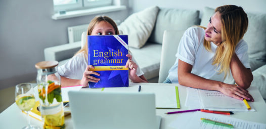 Kind sitzt mit einer Frau an einem Tisch vor einem Laptop und hält ein englisches Grammatikbuch vor das Gesicht; auf dem Tisch liegen Unterlagen, Stifte und ein Getränk. Die Szene wirkt wie gemeinsames Lernen zu Hause.