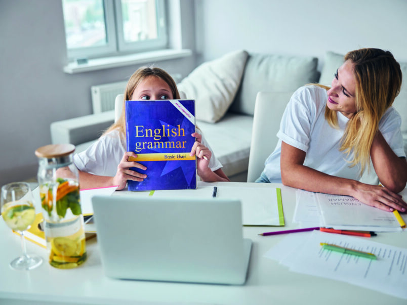 Kind sitzt mit einer Frau an einem Tisch vor einem Laptop und hält ein englisches Grammatikbuch vor das Gesicht; auf dem Tisch liegen Unterlagen, Stifte und ein Getränk. Die Szene wirkt wie gemeinsames Lernen zu Hause.