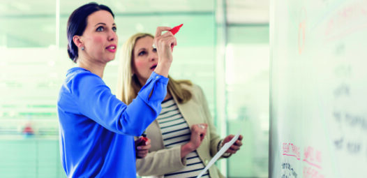 Zwei Frauen stehen vor einem Whiteboard in einem modernen Büro; eine schreibt mit rotem Marker, die andere hält ein Blatt Papier und schaut auf die Notizen.