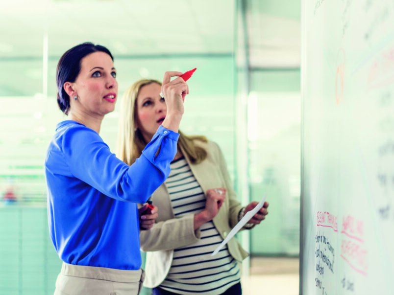 Zwei Frauen stehen vor einem Whiteboard in einem modernen Büro; eine schreibt mit rotem Marker, die andere hält ein Blatt Papier und schaut auf die Notizen.