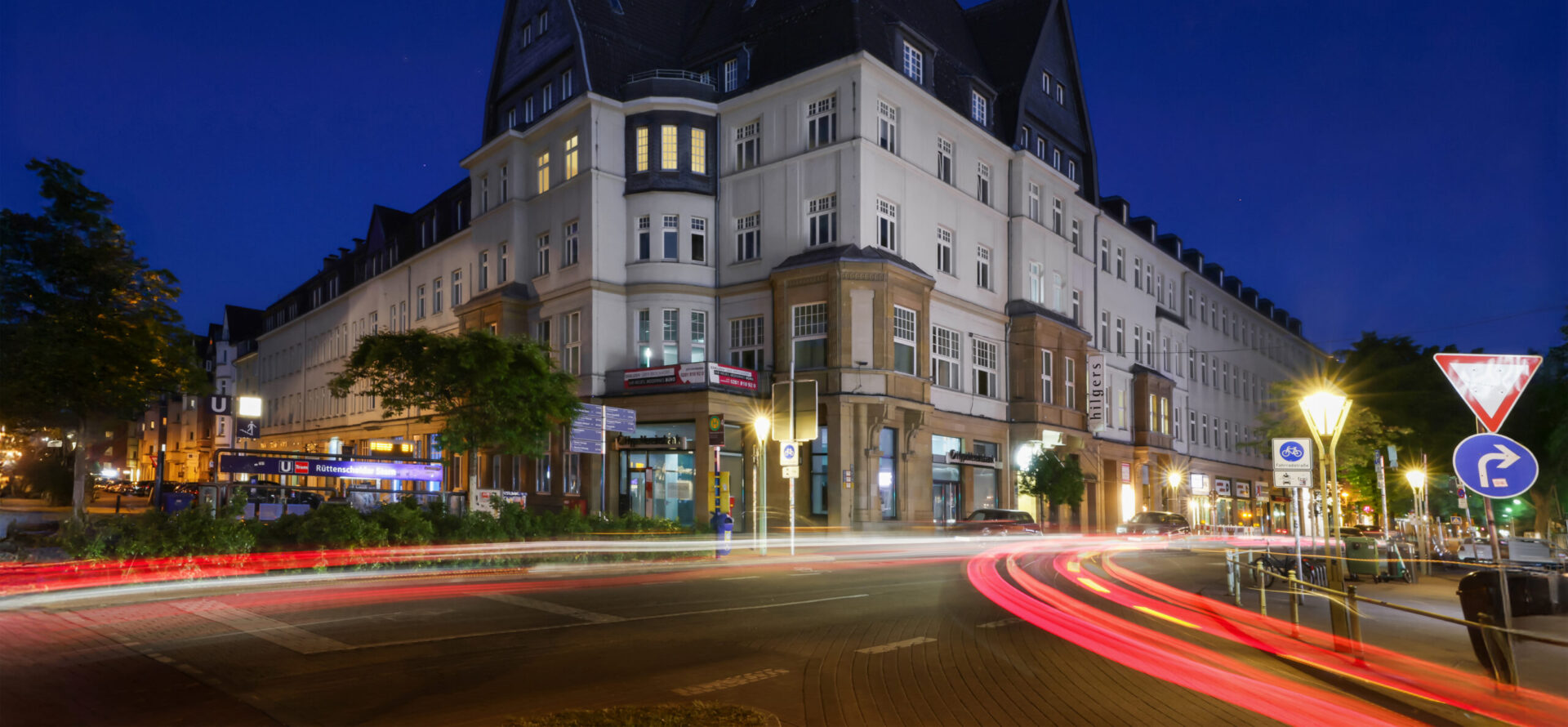 Großes Eckgebäude an einer beleuchteten Straßenkreuzung bei Nacht; die Fenster sind teilweise erhellt, und vorbeifahrende Autos hinterlassen rote und weiße Lichtspuren.