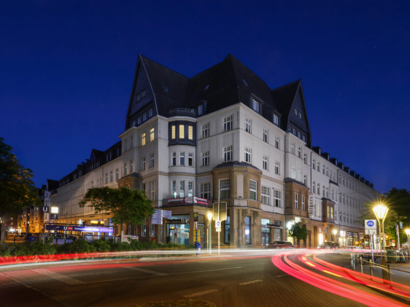 Großes Eckgebäude an einer beleuchteten Straßenkreuzung bei Nacht; die Fenster sind teilweise erhellt, und vorbeifahrende Autos hinterlassen rote und weiße Lichtspuren.