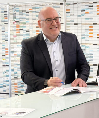 Ein Mann im Anzug steht hinter einem Empfangstresen, lächelt und spricht mit einer Frau; vor ihnen liegen Unterlagen, im Hintergrund ist ein großer Wandplan zu sehen.